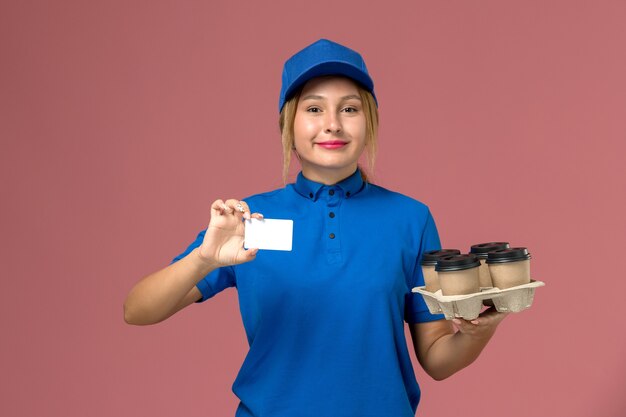 Mensajero femenino en uniforme azul con tarjeta blanca y tazas de café de entrega marrón en rosa, trabajador de entrega uniforme de trabajo de servicio