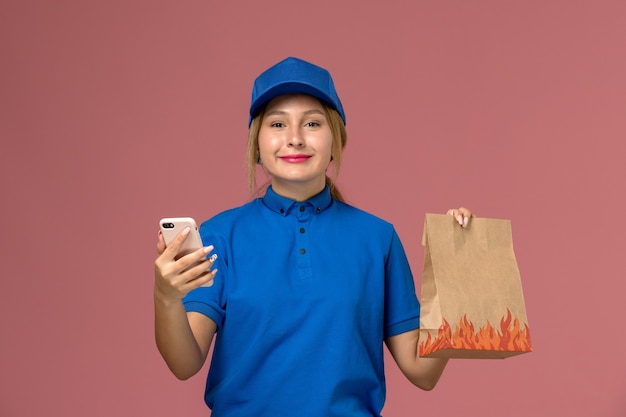 Mensajero femenino en uniforme azul usando su teléfono sosteniendo el paquete de alimentos con una leve sonrisa en rosa claro, trabajo de entrega uniforme de servicio