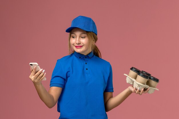 Mensajero femenino en uniforme azul usando su teléfono y sosteniendo tazas de café marrón en rosa claro, entrega uniforme del servicio de trabajo