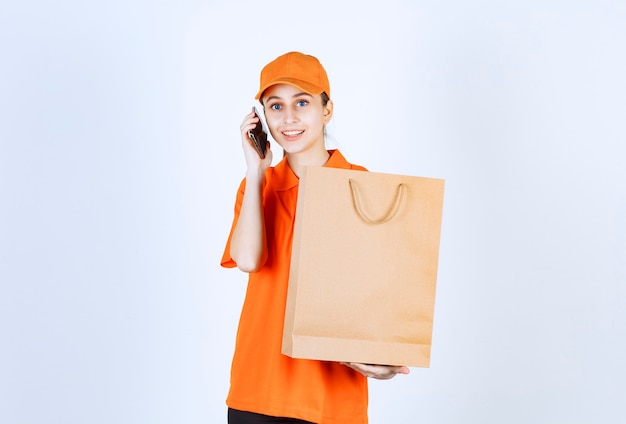 Mensajero femenino en uniforme naranja entregando una bolsa de compras y hablando por teléfono