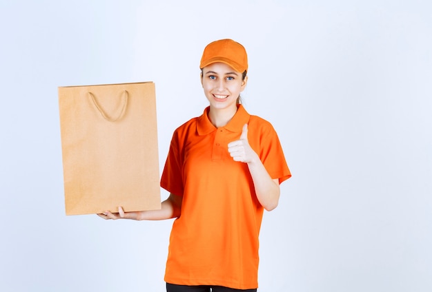 Mensajero femenino en uniforme naranja entregando una bolsa de compras y mostrando un signo de mano positivo