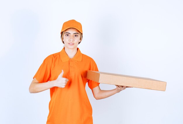 Mensajero femenino en uniforme naranja entregando una caja de cartón y mostrando el signo de la mano de satisfacción