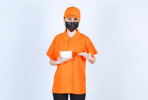 Mensajero femenino en uniforme naranja y máscara negra sosteniendo un vaso de plástico y apuntando a algo