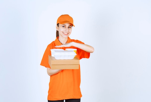 Mensajero femenino en uniforme naranja sosteniendo una caja de cartón, una caja de plástico para llevar y una taza de fideos amarilla