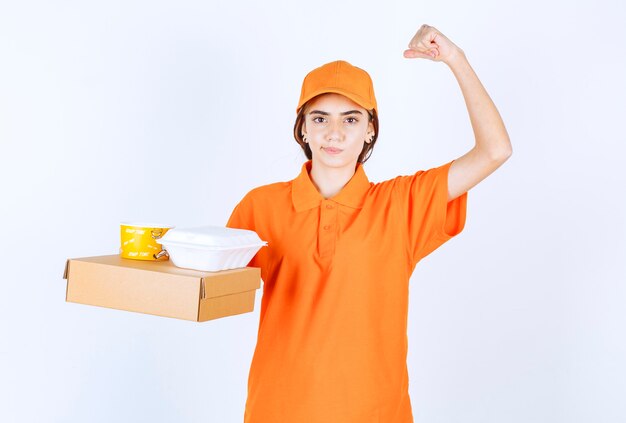 Mensajero femenino en uniforme naranja sosteniendo cajas de comida para llevar amarillas y blancas con un paquete de cartón y disfrutando del sabor