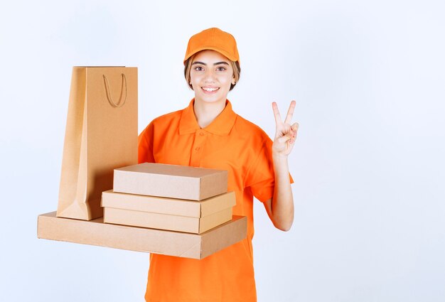 Mensajero femenino en uniforme naranja sosteniendo un stock de paquetes de cartón y bolsas de la compra y mostrando un signo de mano positivo