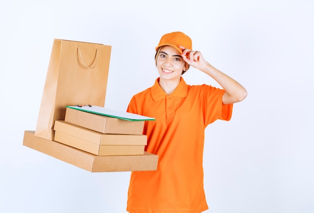 Mensajero femenino en uniforme naranja sosteniendo un stock de paquetes de cartón y bolsas de la compra.