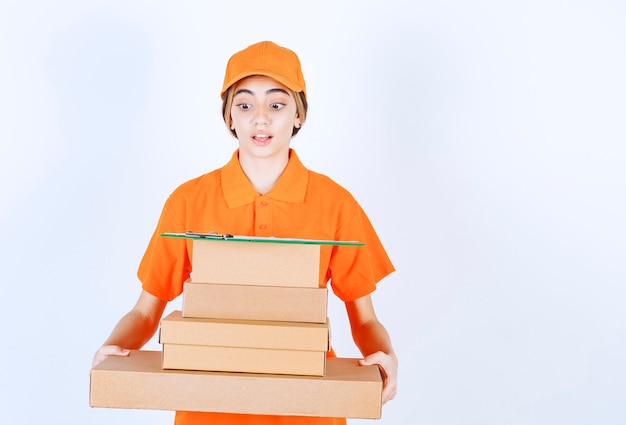Mensajero femenino en uniforme naranja sosteniendo un stock de paquetes de cartón y parece confundido y pensativo