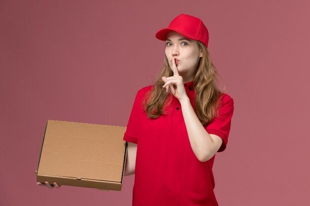 Mensajero femenino en uniforme rojo con caja de comida que muestra el signo de silencio en la entrega del trabajador de servicio uniforme de trabajo de color rosa claro