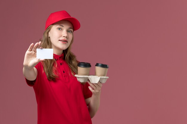 Mensajero femenino en uniforme rojo con entrega tazas de café tarjeta blanca en rosa claro, servicio uniforme de trabajo entrega del trabajador