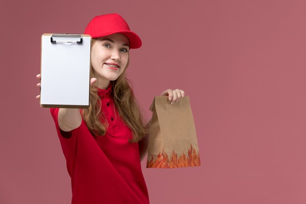 Mensajero femenino en uniforme rojo con paquete de comida marrón con bloc de notas en rosa, servicio de trabajador uniforme entrega