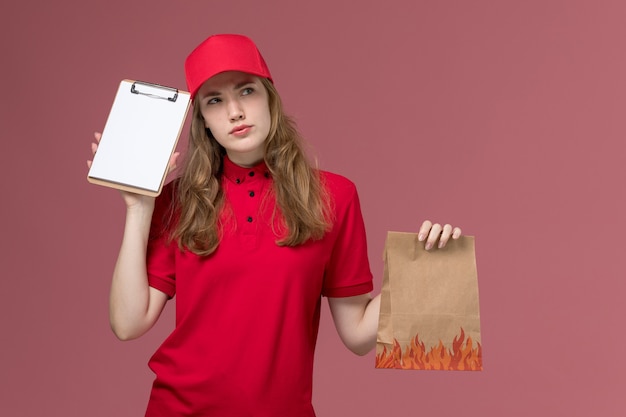 Mensajero femenino en uniforme rojo sosteniendo el bloc de notas y el paquete de alimentos pensando en rosa, uniforme trabajador de trabajo de entrega