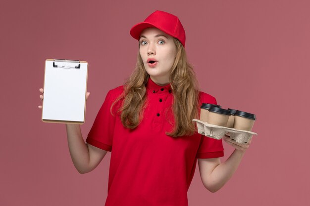 Mensajero femenino en uniforme rojo sosteniendo bloc de notas y tazas de café en rosa, trabajo de trabajador de entrega de servicio uniforme