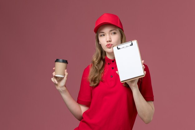 Mensajero femenino en uniforme rojo sosteniendo café y bloc de notas en rosa, servicio de trabajador de trabajo uniforme entrega