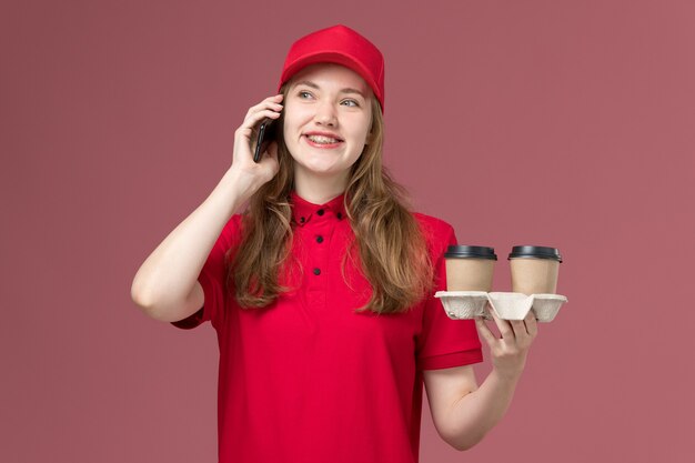 Mensajero femenino en uniforme rojo sosteniendo café de entrega hablando por teléfono en rosa, trabajo de trabajador de entrega de servicio uniforme