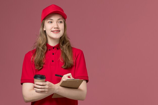Mensajero femenino en uniforme rojo sosteniendo café escribiendo notas en rosa claro, servicio de entrega uniforme de trabajo trabajador