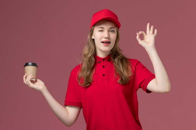 Mensajero femenino en uniforme rojo sosteniendo la taza de café guiñando un ojo en rosa, trabajador de trabajo de entrega de servicio uniforme