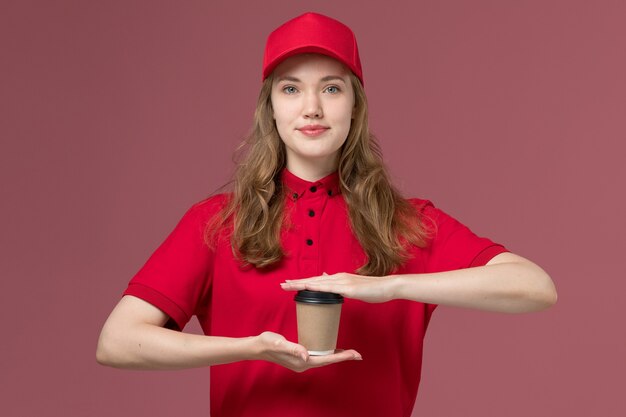 Mensajero femenino en uniforme rojo sosteniendo una taza de café marrón en rosa, trabajo de trabajador de entrega de servicio uniforme