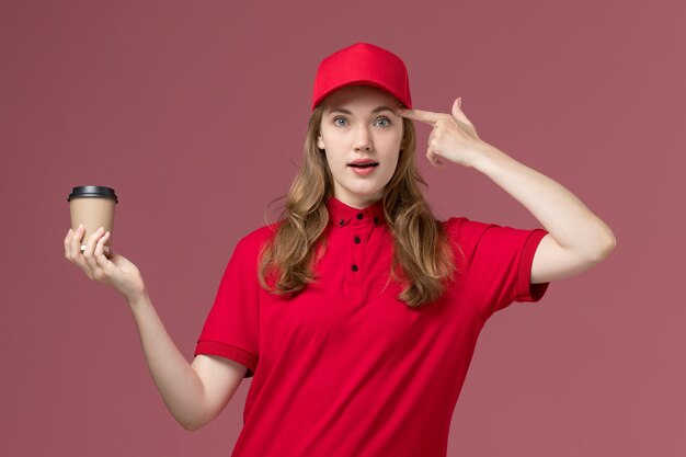 Mensajero femenino en uniforme rojo sosteniendo la taza de café posando en rosa claro, entrega de trabajador de servicio uniforme de trabajo