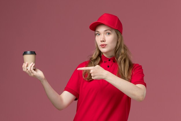 Mensajero femenino en uniforme rojo sosteniendo la taza de café posando en rosa claro, entrega de trabajador de servicio uniforme de trabajo