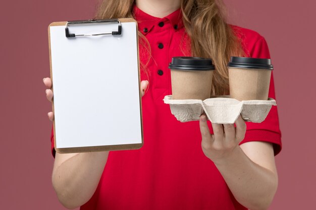 Mensajero femenino en uniforme rojo sosteniendo tazas de café de bloc de notas en rosa, trabajador de entrega de servicio uniforme