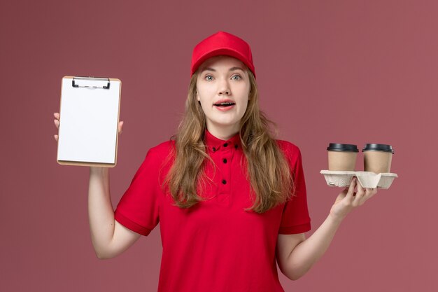 Mensajero femenino en uniforme rojo sosteniendo tazas de café y bloc de notas en rosa, trabajo de entrega de servicio uniforme