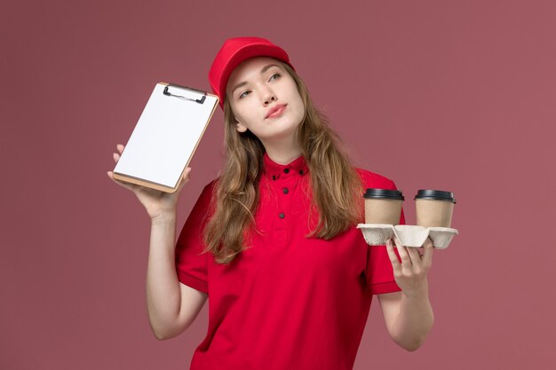 Mensajero femenino en uniforme rojo sosteniendo tazas de café con bloc de notas sonriendo en rosa, trabajador de entrega de servicio uniforme