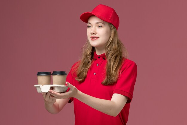 Mensajero femenino en uniforme rojo sosteniendo tazas de café de entrega entregándolas en rosa claro, servicio de trabajo uniforme trabajador repartidor