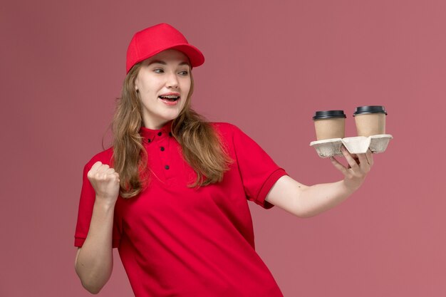 Mensajero femenino en uniforme rojo sosteniendo tazas de café de entrega marrón en el trabajador de trabajo de entrega de servicio uniforme rosa