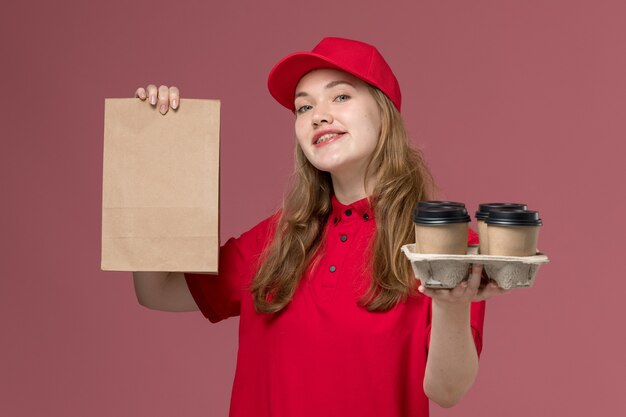 Mensajero femenino en uniforme rojo sosteniendo tazas de café de entrega y paquete de comida en rosa claro, servicio de trabajo uniforme chica de entrega del trabajador