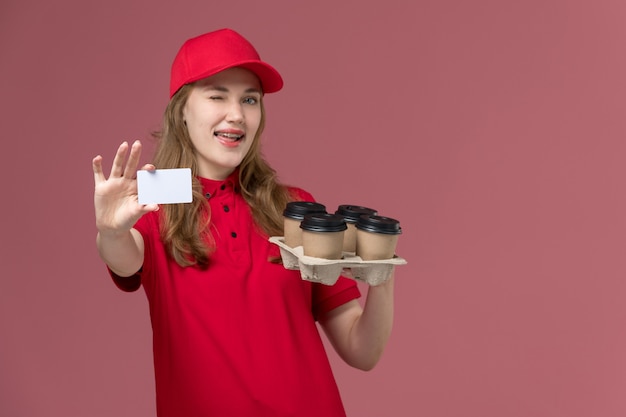 Mensajero femenino en uniforme rojo sosteniendo tazas de café de entrega y tarjeta de plástico blanco en rosa claro, servicio de trabajo uniforme chica de entrega