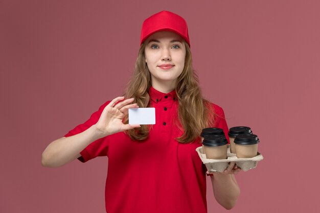 Mensajero femenino en uniforme rojo sosteniendo tazas de café de entrega y tarjeta de plástico en rosa claro, servicio de trabajo uniforme trabajador repartidor