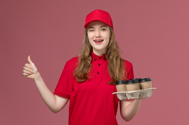 Mensajero femenino en uniforme rojo sosteniendo tazas de café guiñando un ojo en rosa, servicio de trabajador de trabajo uniforme