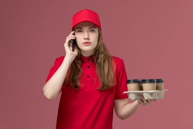 Mensajero femenino en uniforme rojo sosteniendo tazas de café hablando por teléfono en rosa claro, entrega de servicio de trabajador uniforme de trabajo