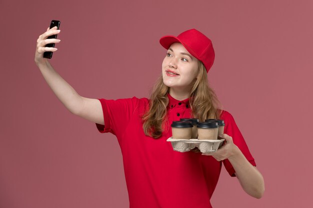 Mensajero femenino en uniforme rojo sosteniendo tazas de café marrón tomando selfie en rosa, trabajo de trabajador de entrega de servicio uniforme