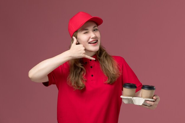 Mensajero femenino en uniforme rojo sosteniendo tazas de café y posando guiñando un ojo en el trabajo de trabajador de entrega de servicio uniforme rosa