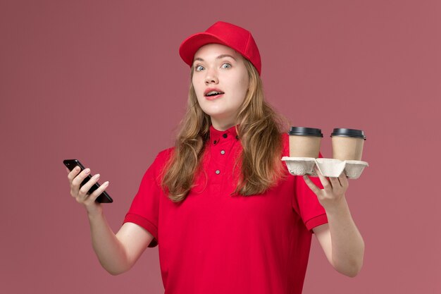 Mensajero femenino en uniforme rojo sosteniendo tazas de café y teléfono inteligente en rosa, trabajo de entrega de servicio uniforme