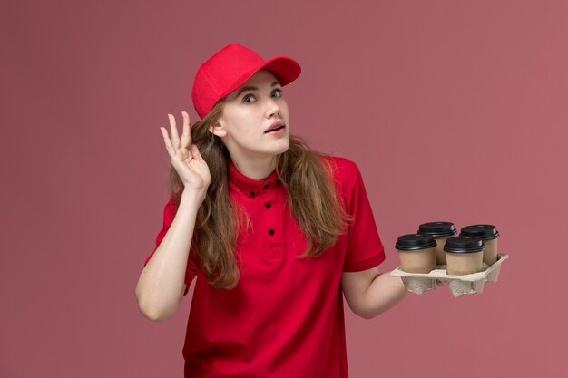 Mensajero femenino en uniforme rojo sosteniendo tazas de café tratando de escuchar sobre rosa, trabajador de trabajo de entrega de servicio uniforme