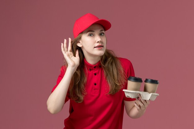 Mensajero femenino en uniforme rojo sosteniendo tazas de café y tratando de escuchar sobre el trabajo de trabajador de entrega de servicio uniforme rosa