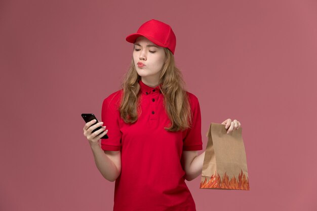 Mensajero femenino en uniforme rojo sosteniendo el teléfono y el paquete de alimentos en rosa claro, servicio de entrega uniforme del trabajador