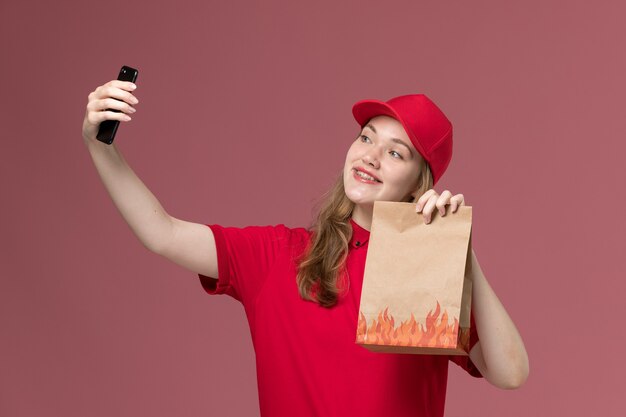 Mensajero femenino en uniforme rojo tomando selfie sosteniendo el paquete de comida de papel en rosa claro, servicio uniforme de trabajo entrega de trabajador