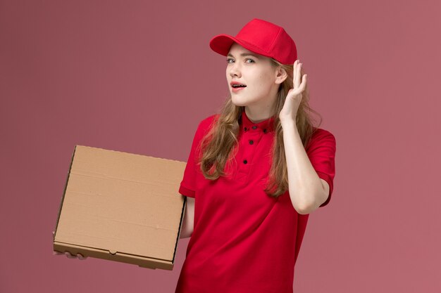 Mensajero femenino en uniforme rojo tratando de escuchar sosteniendo la caja de comida en rosa, trabajador de trabajo de entrega de servicio uniforme