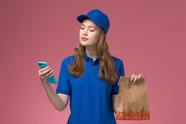 Mensajero femenino de vista frontal en azul sosteniendo un paquete de teléfono y comida en el trabajo de la empresa uniforme de servicio de escritorio de luz rosa