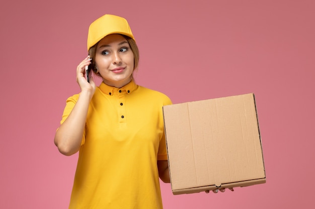 Foto gratuita mensajero femenino de vista frontal en capa amarilla uniforme amarillo hablando por teléfono sosteniendo el paquete de entrega en el trabajo de entrega uniforme de fondo rosa