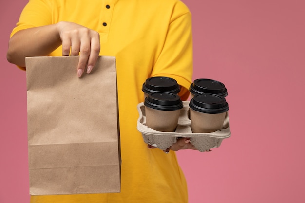Mensajero femenino de vista frontal en capa amarilla uniforme amarillo con paquete de alimentos y café en el color de trabajo de trabajo de entrega uniforme de fondo rosa