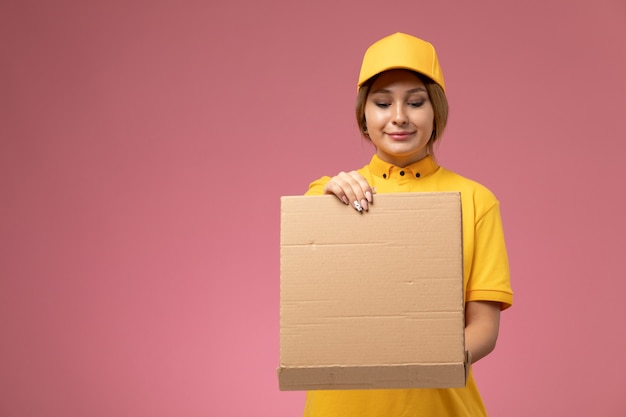 Mensajero femenino de vista frontal en capa amarilla uniforme amarillo sosteniendo y abriendo la caja de comida en el color femenino de entrega uniforme de escritorio rosa