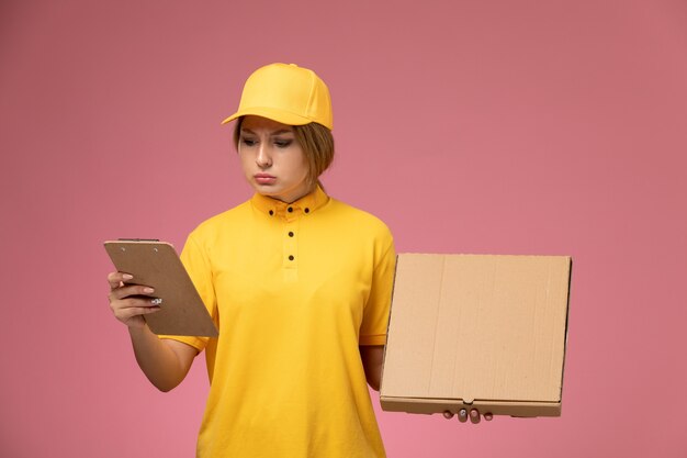 Mensajero femenino de vista frontal en capa amarilla uniforme amarillo sosteniendo el bloc de notas de caja de comida en el escritorio rosa uniforme femenino de entrega