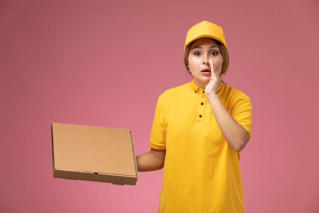 Mensajero femenino de vista frontal en capa amarilla uniforme amarillo sosteniendo la caja de comida y susurrando en el color femenino de entrega uniforme de escritorio rosa