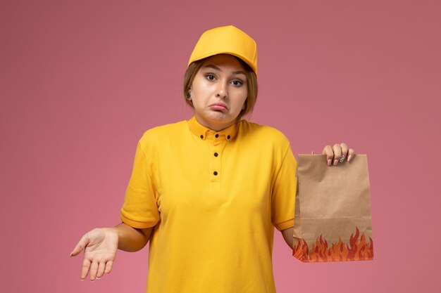 Mensajero femenino de vista frontal en capa amarilla uniforme amarillo sosteniendo el paquete de alimentos en el color de trabajo de entrega uniforme de fondo rosa