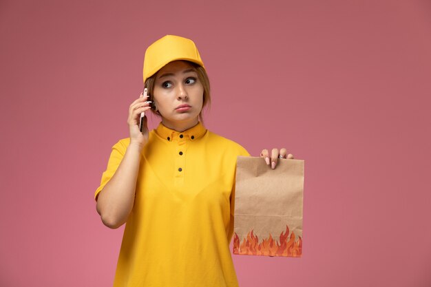 Mensajero femenino de vista frontal en capa amarilla uniforme amarillo sosteniendo el paquete de alimentos hablando por teléfono sobre fondo rosa trabajo de entrega uniforme trabajo de color